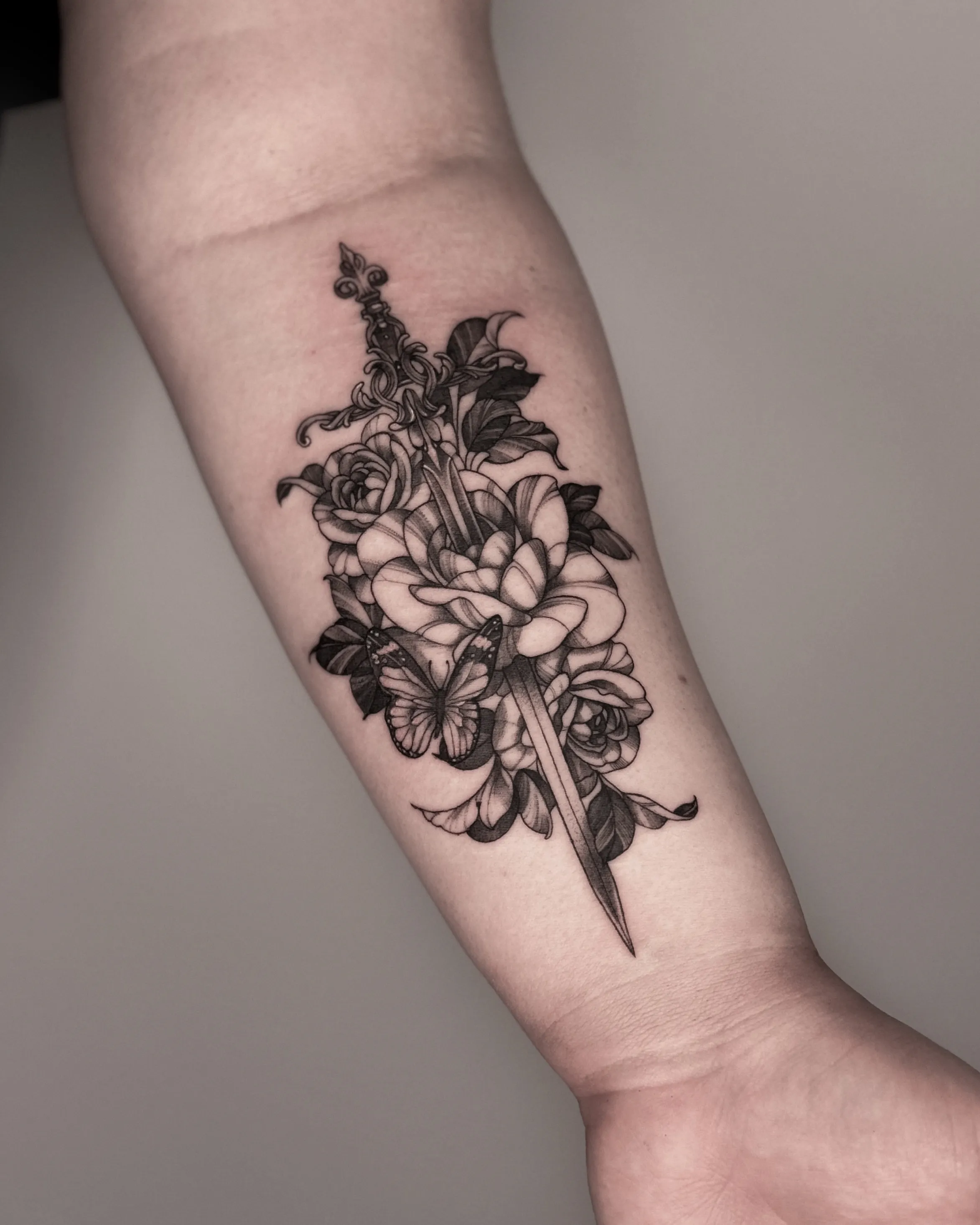 Black and gray tattoo of a dagger with roses and a butterfly on the forearm